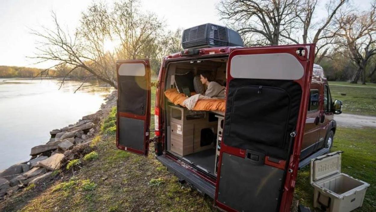 Rear of the Winnebago Solis parked by a lake with back doors open. A woman reads a book inside.