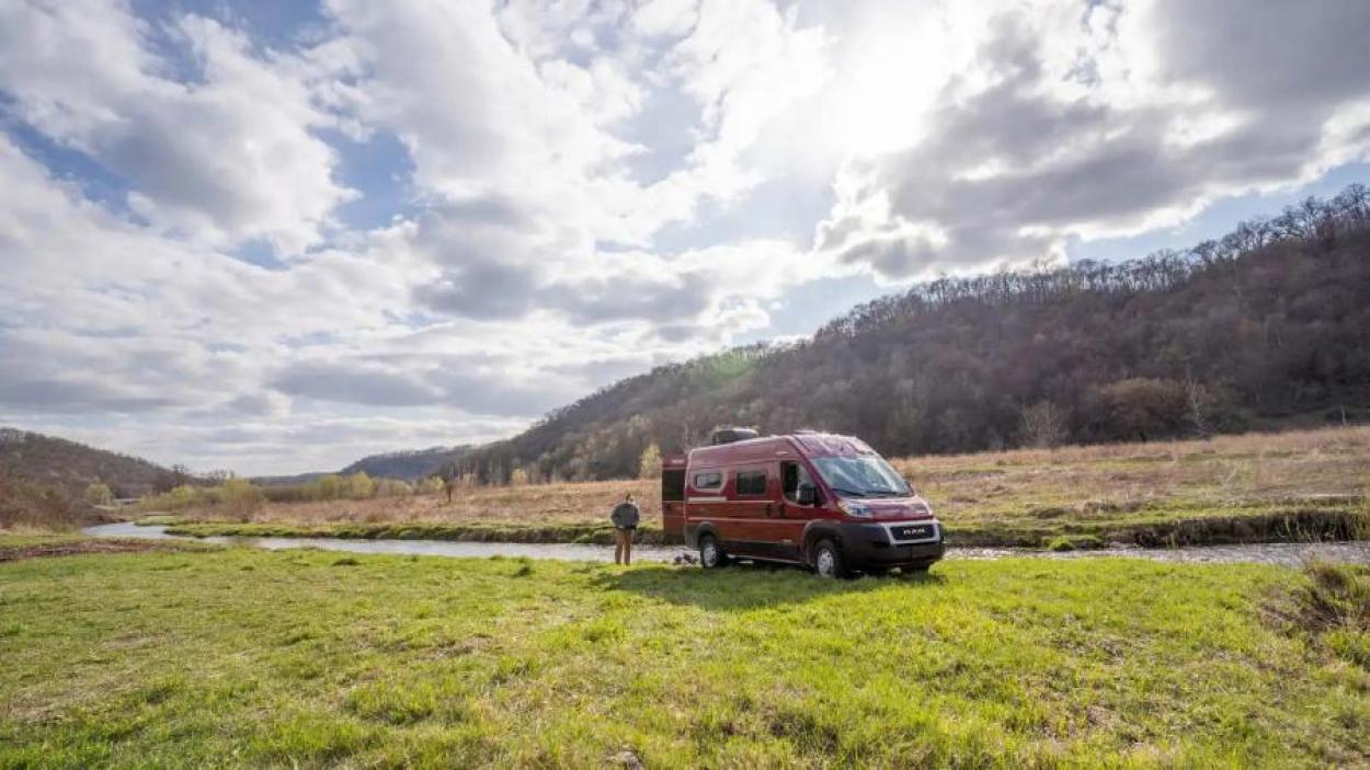 Man getting his fishing gear together behind his red Winnebago Solis campervan, parked by a creek on a grassy plain