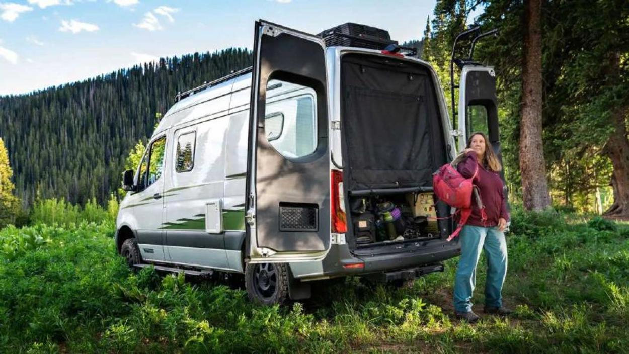 Woman slings her backpack over her shoulder as she stands behind a Winnebago Revel adventure Class B RV