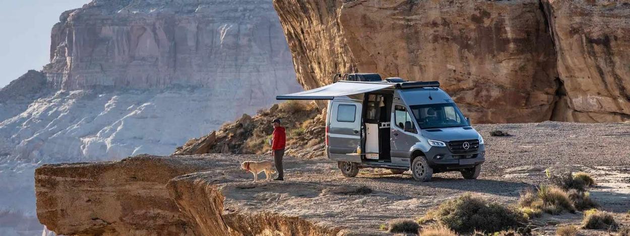 Man and dog standing outside a Winnebago Revel parked on a rocky outcrop, with the campervan's awning outstretched