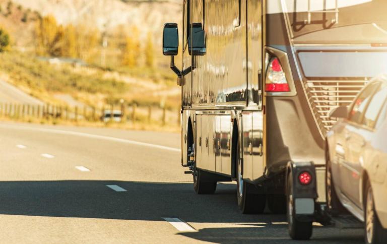 View of the rear of a Class A RV towing a small car during golden hour on the highway