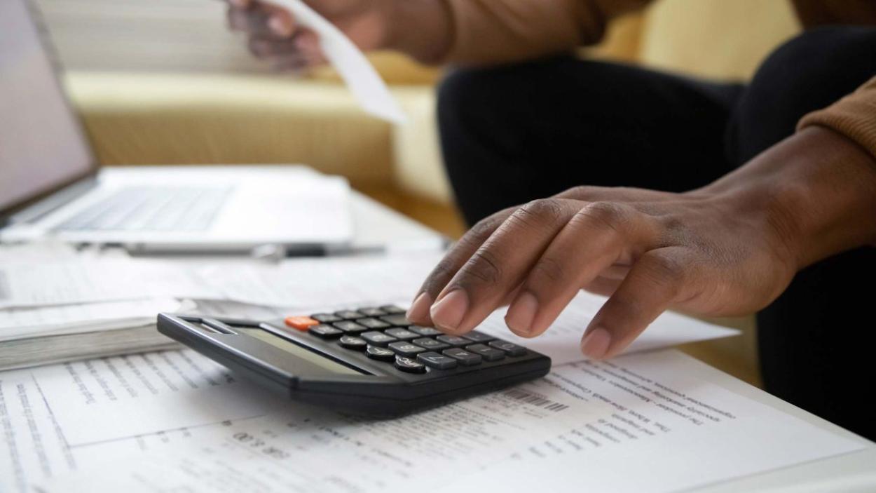 Man&#039;s hand above a calculator with paperwork and a computer in the background